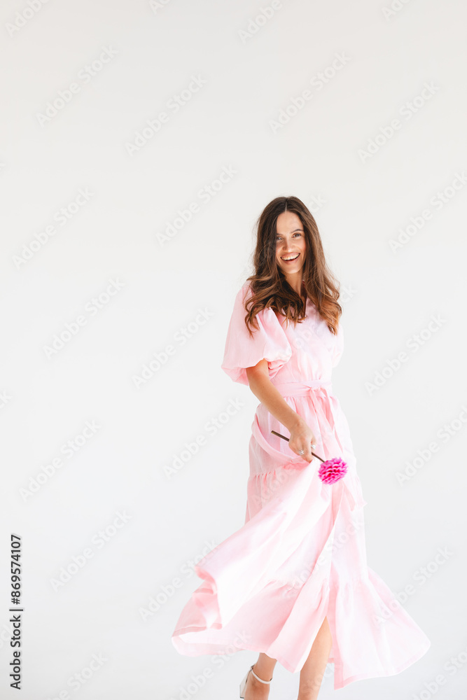 Model wearing casual outfit. Look in trendy minimalistic style. Pink dress with slit, belt, puff sleeves. Sexy sensual happy woman. Positive woman turn around and hold pink flower on white background. Model wearing casual outfit. Look in trendy minimalistic style. Pink dress with slit, belt, puff sleeves. Sexy sensual happy woman. Positive woman turn around and hold pink flower on white background.