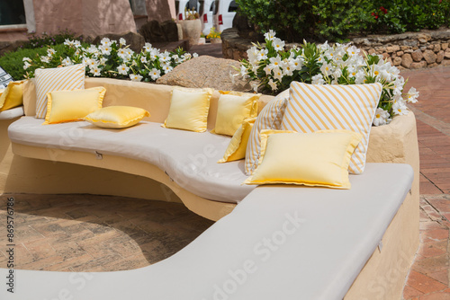 Garden and terrace design or landscaping: Outdoor brick-built bench with white and yellow cushions surrounded by bushes of flourishing white dipladenia or mandevilla flowers