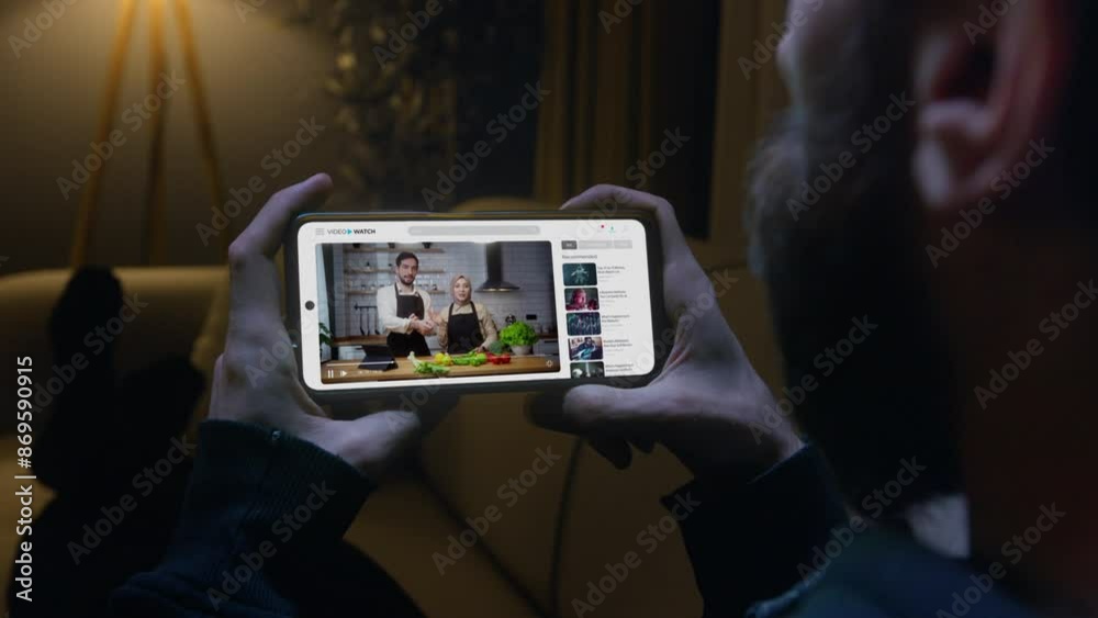 Man laying on sofa watching a cooking tutorial on his smartphone in living room. Modern digital learning and the convenience of accessing educational content online at home