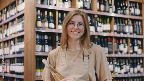 Inside a wine boutique, a cheerful female shopkeeper in an apron warmly assists customers amid a neat and tidy shop. The store offers a wide selection of beverages to choose from