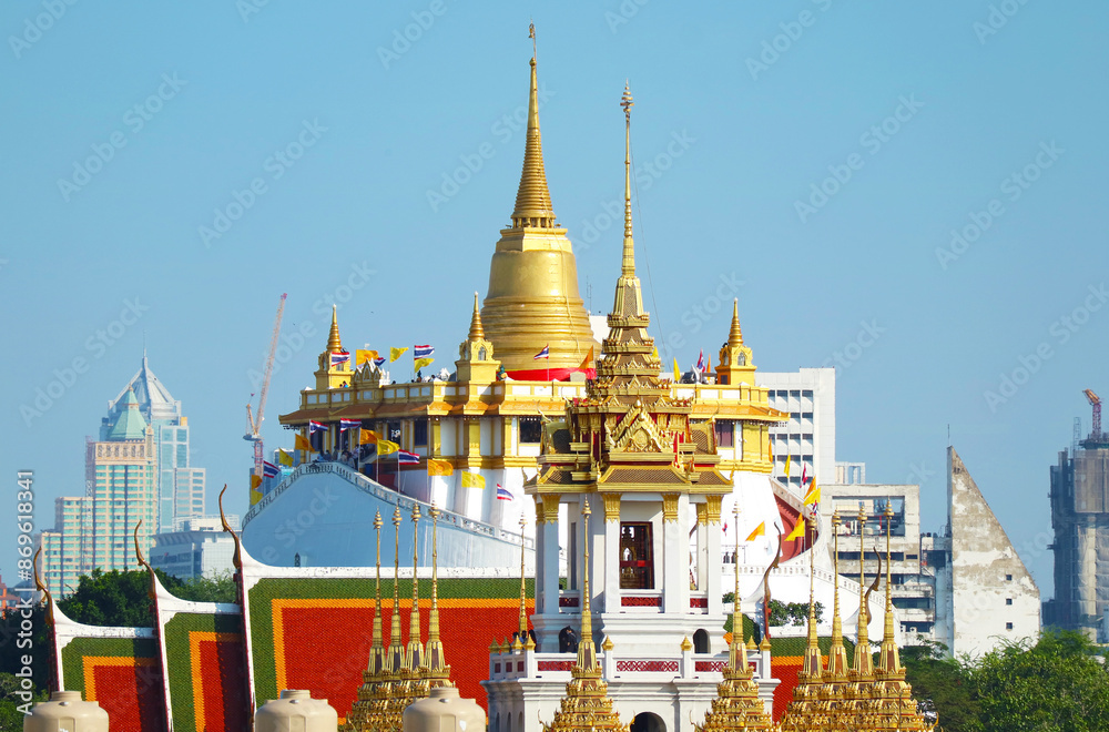 Naklejka premium Stunning Phu Khao Thong (Golden Mount) of Wat Saket Temple with the Spires of Loha Prasat (Iron Castle) of Wat Ratchanatdaram Temple in Foreground, Bangkok City Skyline, Thailand