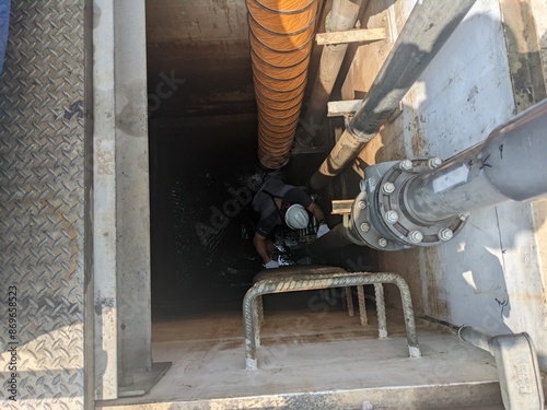 Two Workers Inspecting a Pipe Inside a Concrete Structure Confine space. A worker wearing a safety helmet and ventilation equipment descends into a confined space through a manhole. The space is equip