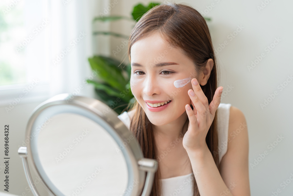 Facial beauty skin health care, smile of cute asian young woman looking at mirror, hand applying moisturizer lotion on her face, female putting cream treatment before makeup cosmetic routine at home. Facial beauty skin health care, smile of cute asian young woman looking at mirror, hand applying moisturizer lotion on her face, female putting cream treatment before makeup cosmetic routine at home.