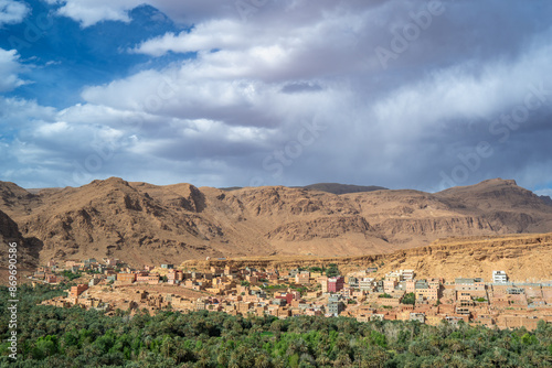 Lush palm trees surround mud-brick buildings against towering cliffs under a dynamic sky. Captured in Todra Gorges, Morocco.