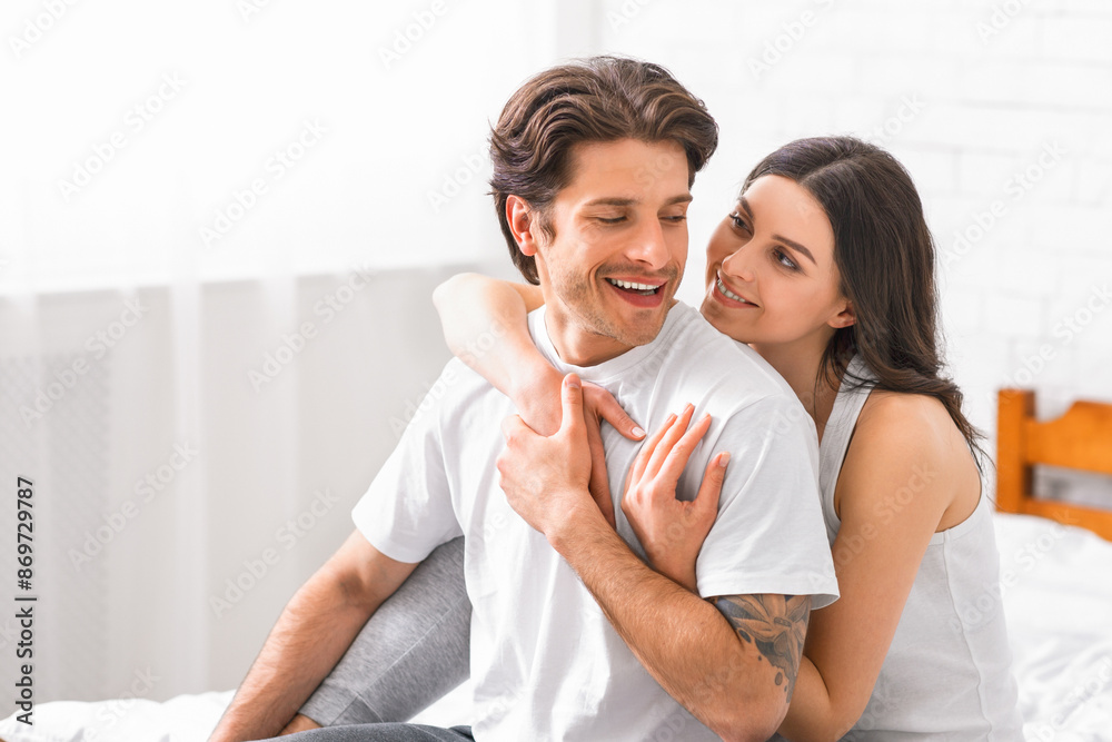 A smiling man sits on a bed with a woman hugging him from behind, both wearing casual clothes in a brightly lit bedroom. A smiling man sits on a bed with a woman hugging him from behind, both wearing casual clothes in a brightly lit bedroom.