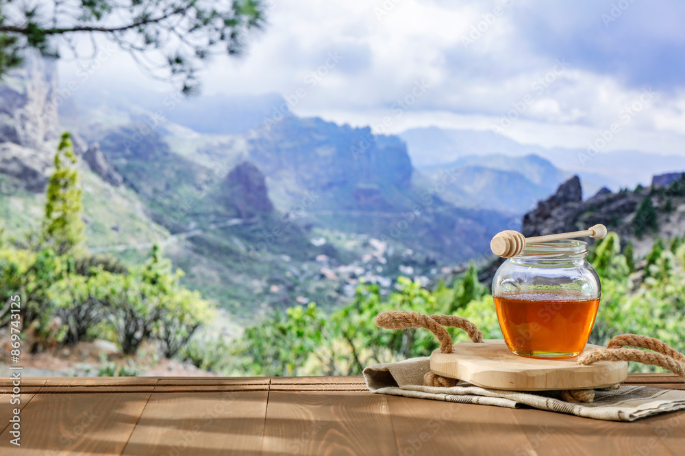 A jar of honey on wooden table top and blur green natural meadow and grass and trees and mountains background. Copy space for food and advertising products. A jar of honey on wooden table top and blur green natural meadow and grass and trees and mountains background. Copy space for food and advertising products.