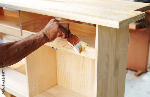 Carpenter hands applying varnish to a cabinet with a paintbrush. First lacquer coating on wood surface at a furniture and wooden workshop.Close up.