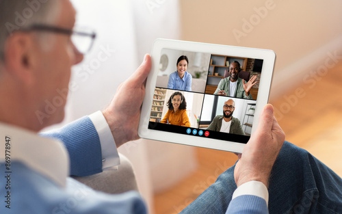 An older man wearing eyeglasses is sitting on a couch, holding a tablet and participating in a video conference with three other colleagues