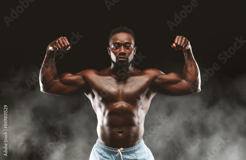 A muscular man flexes his biceps, showcasing his strength and physique against a dark background with subtle smoke effects.