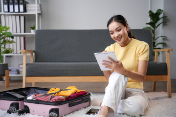 Asian woman sitting on floor at home writing packing list for travel trip