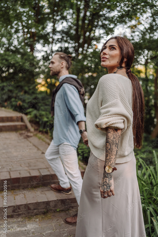 Outdoor shot of young couple in love walking on pathway through grass field. Young couple strolling in the park. Trendy couple with tattoos walking in a park. Botanic Garden. Outdoor shot of young couple in love walking on pathway through grass field. Young couple strolling in the park. Trendy couple with tattoos walking in a park. Botanic Garden.