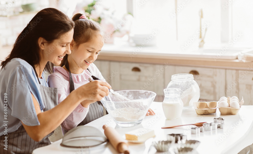 A mother and daughter are baking together in a bright kitchen. The daughter is stirring a bowl of flour with a whisk, while the mother watches with a smile. They are both wearing aprons A mother and daughter are baking together in a bright kitchen. The daughter is stirring a bowl of flour with a whisk, while the mother watches with a smile. They are both wearing aprons