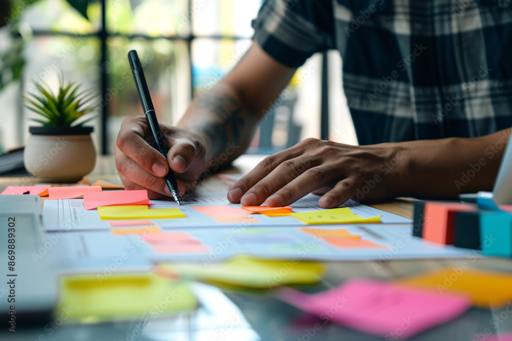 Person writing on paper with colorful sticky notes on a desk in a creative workspace setting Person writing on paper with colorful sticky notes on a desk in a creative workspace setting