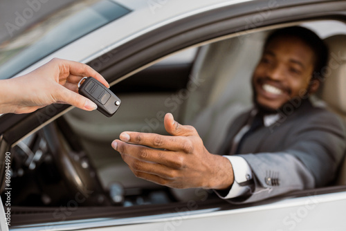 African American man is sitting in the drivers seat of a car with a person handing him the keys. He is smiling and reaching out to take them. © Prostock-studio