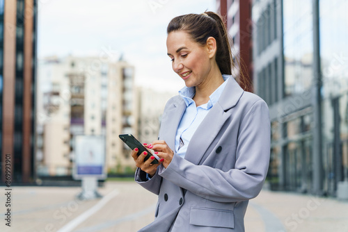 A businesswoman in a grey suit smiles while using her phone, standing in an urban area with modern buildings. © muse studio