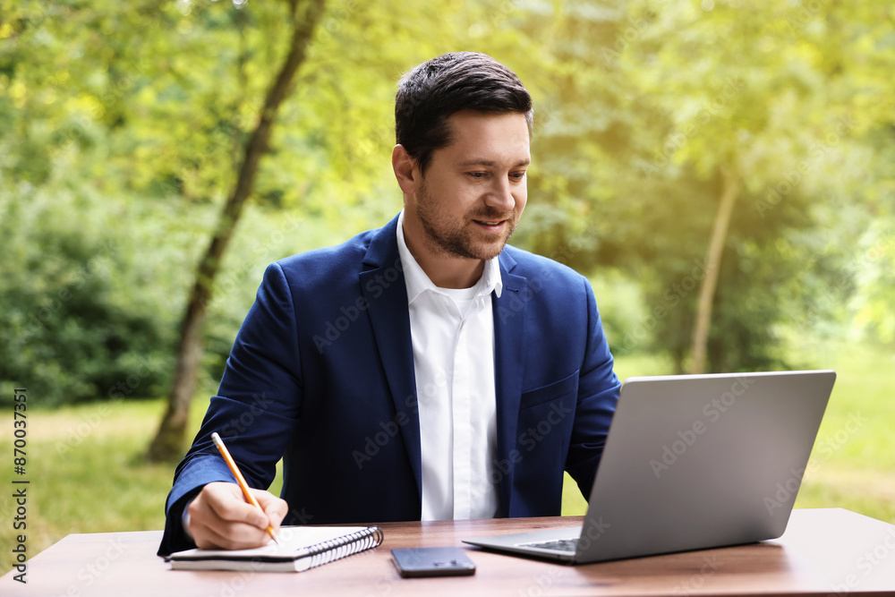 Smiling businessman working with laptop and writing something at table outdoors. Remote job Smiling businessman working with laptop and writing something at table outdoors. Remote job