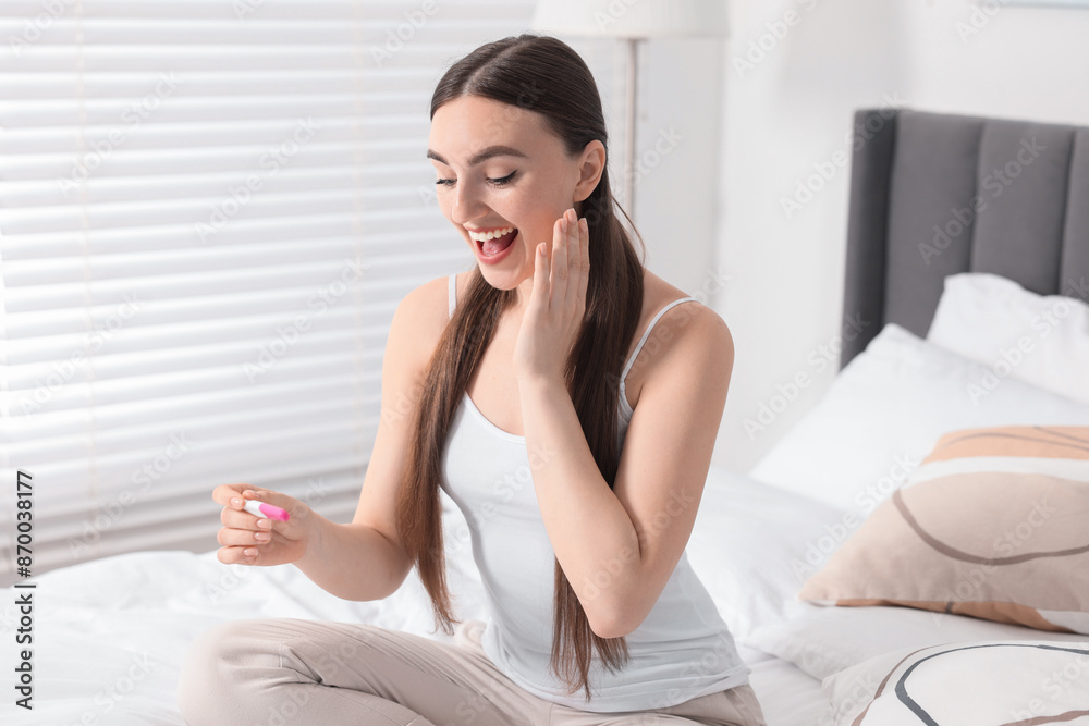 Happy woman holding pregnancy test on bed in room Happy woman holding pregnancy test on bed in room