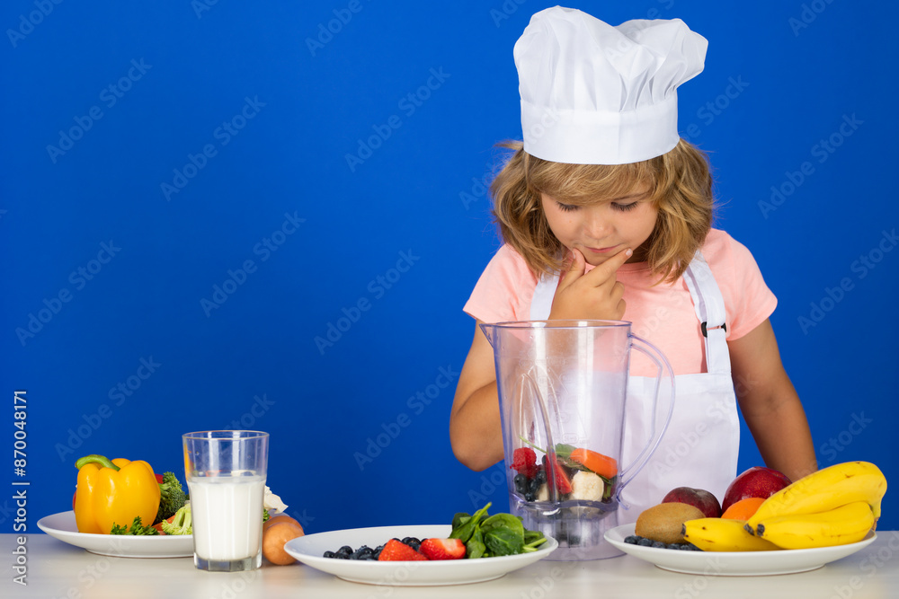 Child chef cook prepares food in isolated blue studio background. Kids cooking. Teen boy with apron and chef hat preparing a healthy vegetables meal in the kitchen. Child chef cook prepares food in isolated blue studio background. Kids cooking. Teen boy with apron and chef hat preparing a healthy vegetables meal in the kitchen.