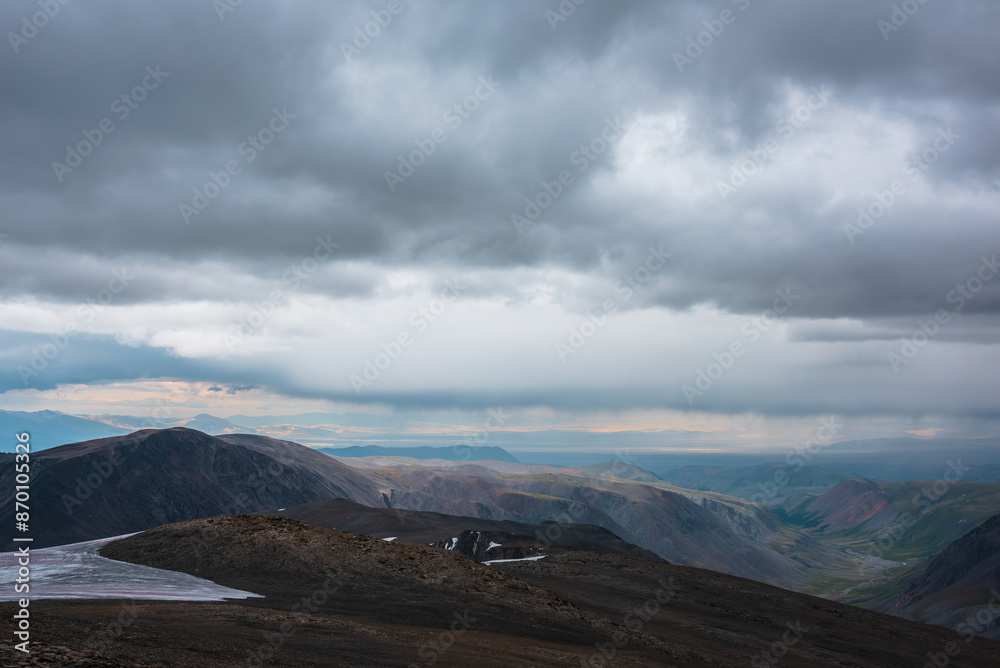 Dramatic top view from precipice edge to wide colorful valley and mountain silhouettes in rain under lead gray cloudy sky. Dark atmospheric mountains in rainy weather and sunset landscape far away. Dramatic top view from precipice edge to wide colorful valley and mountain silhouettes in rain under lead gray cloudy sky. Dark atmospheric mountains in rainy weather and sunset landscape far away.