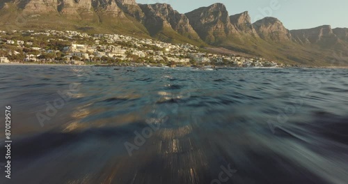 Drone flies fast over the wavy sea at Camps Bay beach in Cape Town South Africa - many houses on a hilltop - view of table mountain rocks in the sea - drone flies through bird swarm