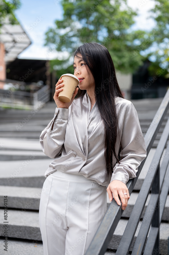 An attractive Asian businesswoman is having her coffee while standing on the stairs outdoors. An attractive Asian businesswoman is having her coffee while standing on the stairs outdoors.