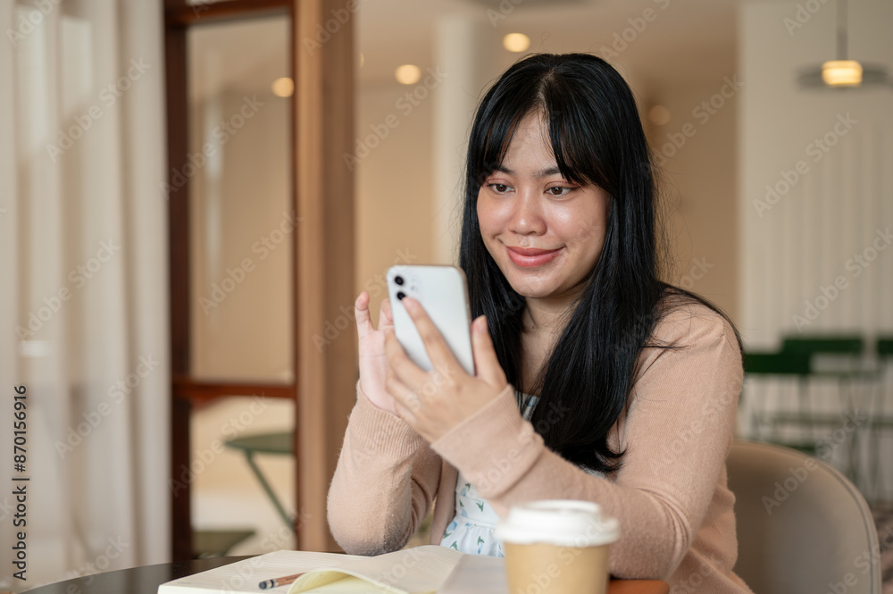 A charming Asian woman is using her smartphone while sitting in a coffee shop with her books. A charming Asian woman is using her smartphone while sitting in a coffee shop with her books.