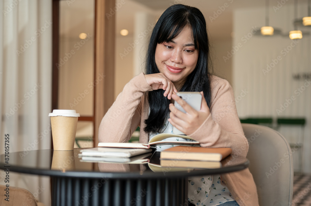 A happy, beautiful Asian woman using her smartphone at a table indoor while reading books. A happy, beautiful Asian woman using her smartphone at a table indoor while reading books.
