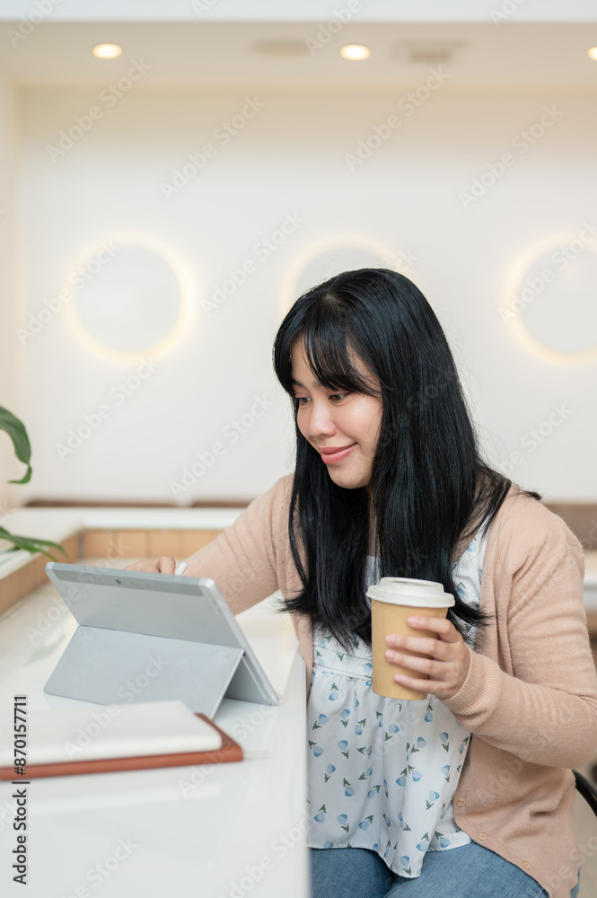 An attractive Asian woman having a hot drink and using her digital tablet in a minimalist cafe. An attractive Asian woman having a hot drink and using her digital tablet in a minimalist cafe.