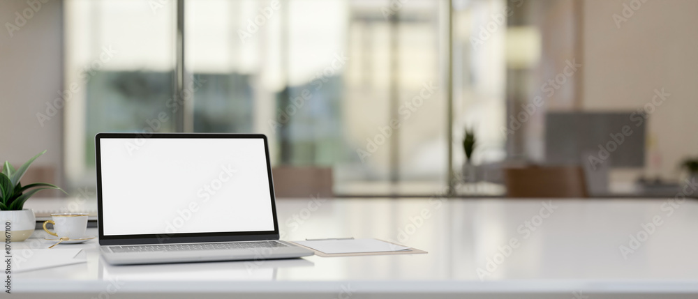 A laptop computer with a white-screen mockup on a table in a modern office. A laptop computer with a white-screen mockup on a table in a modern office.