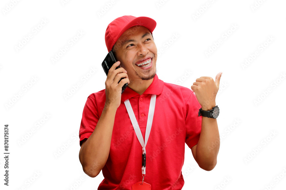 An excited Asian delivery man or courier pointing with thumb at copy space while having an engaging phone call. Isolated on a white background An excited Asian delivery man or courier pointing with thumb at copy space while having an engaging phone call. Isolated on a white background