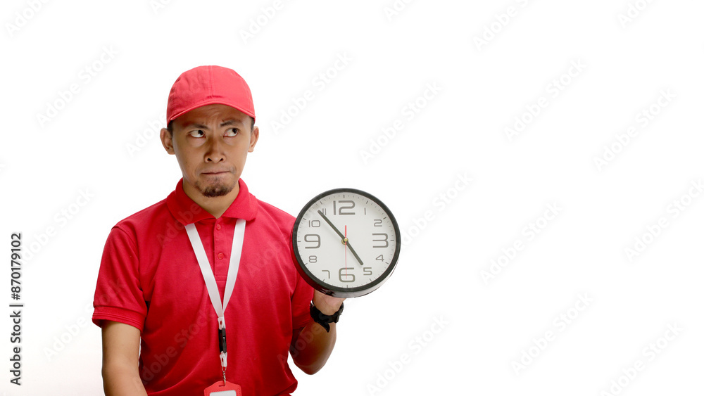Confused Asian delivery man or courier holds a wall clock while looking at an empty copy space, isolated on white background. Concept of time management, deadlines, or scheduling in delivery services Confused Asian delivery man or courier holds a wall clock while looking at an empty copy space, isolated on white background. Concept of time management, deadlines, or scheduling in delivery services