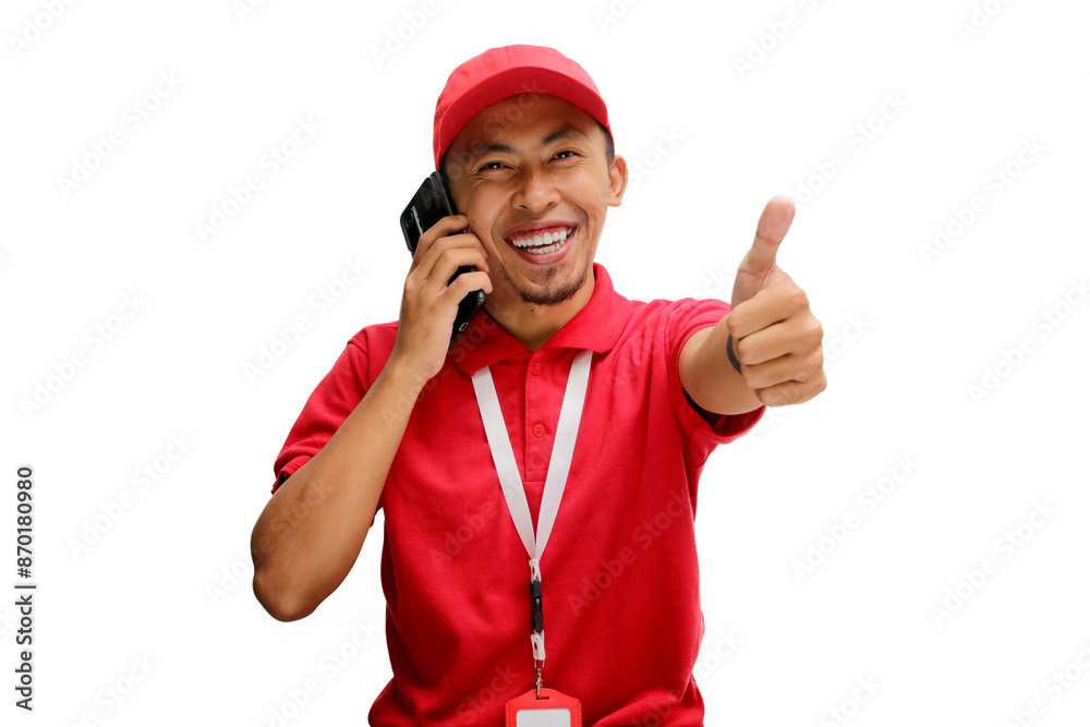Excited Asian delivery man or courier showing a thumbs up gesture to the camera while on a phone call. Isolated on a white background