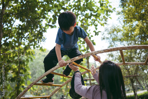 Fotografie A young 8 years old boy climbing a jungle gym with the assistance of his mother in a park, surrounded by green trees and natural light