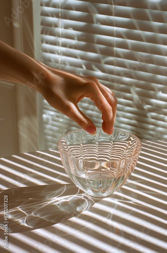 Hand Reaching for Glass Bowl With Water on Table in Sunlight Through Blinds
