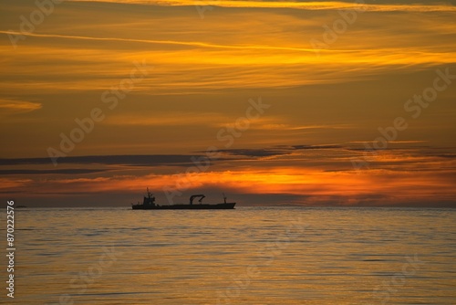 Silhouette of a cargo ship against a vibrant orange sunset over calm ocean waters