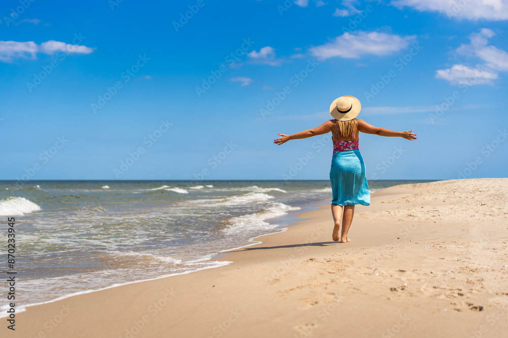 Summer vacation. Beautiful young woman in colorful swimsuit and blue pareo and hat walking with her arms spread out on the sandy beach on a beautiful sunny day. Back view Summer vacation. Beautiful young woman in colorful swimsuit and blue pareo and hat walking with her arms spread out on the sandy beach on a beautiful sunny day. Back view