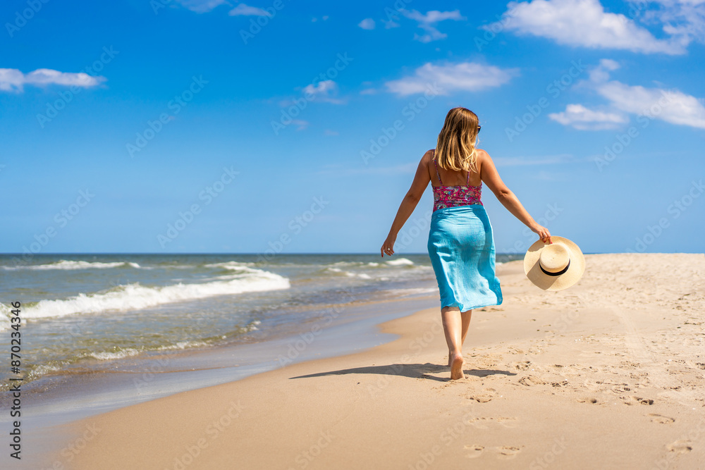 Summer vacation. Beautiful young woman in colorful swimsuit and blue pareo walking with hat in hand on the sandy beach on a beautiful sunny day. Back view Summer vacation. Beautiful young woman in colorful swimsuit and blue pareo walking with hat in hand on the sandy beach on a beautiful sunny day. Back view
