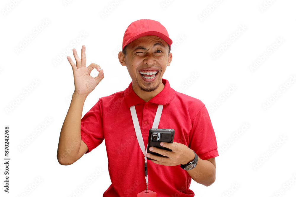 Asian delivery man or courier smiling and showing the OK gesture and winking to the camera while holding a phone. Isolated on a white background Asian delivery man or courier smiling and showing the OK gesture and winking to the camera while holding a phone. Isolated on a white background