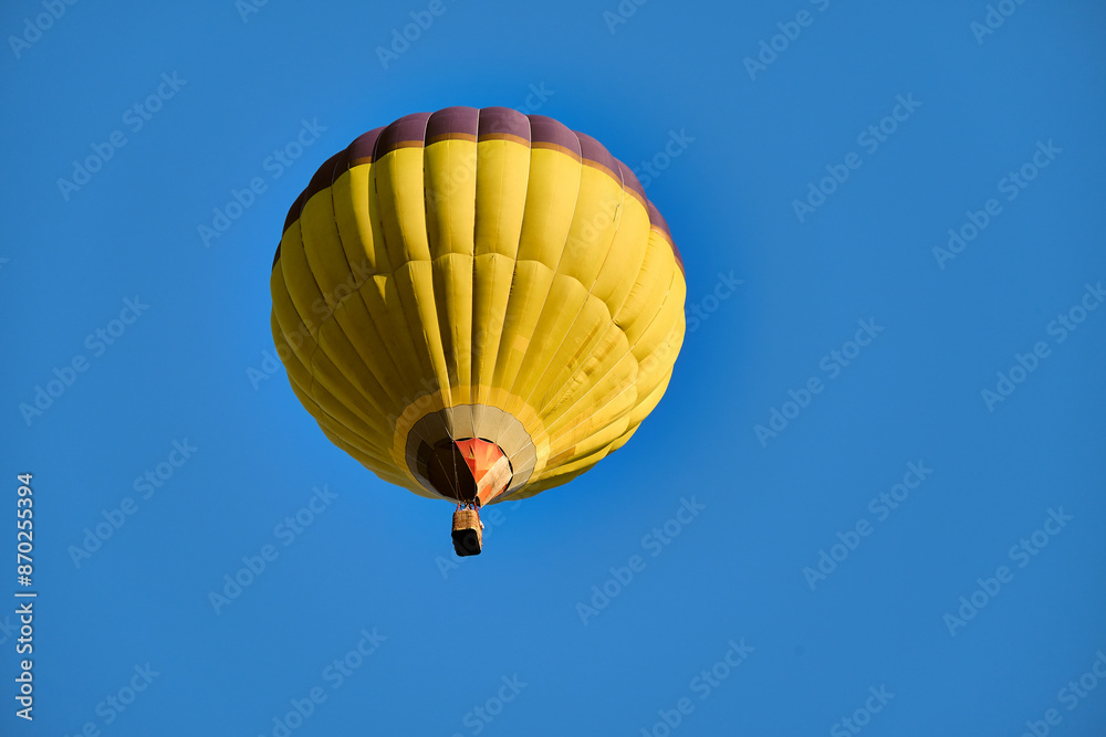 Naklejka premium A vibrant yellow hot air balloon floats gracefully in a clear blue sky, offering a sense of adventure and freedom.