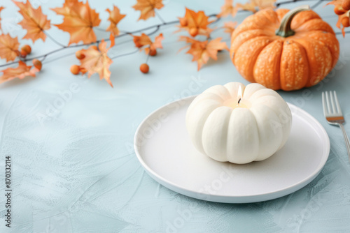 A white pumpkin candle sits on a white plate in the center of a Thanksgiving table setting, decorated with fall leaves and a single orange pumpkin