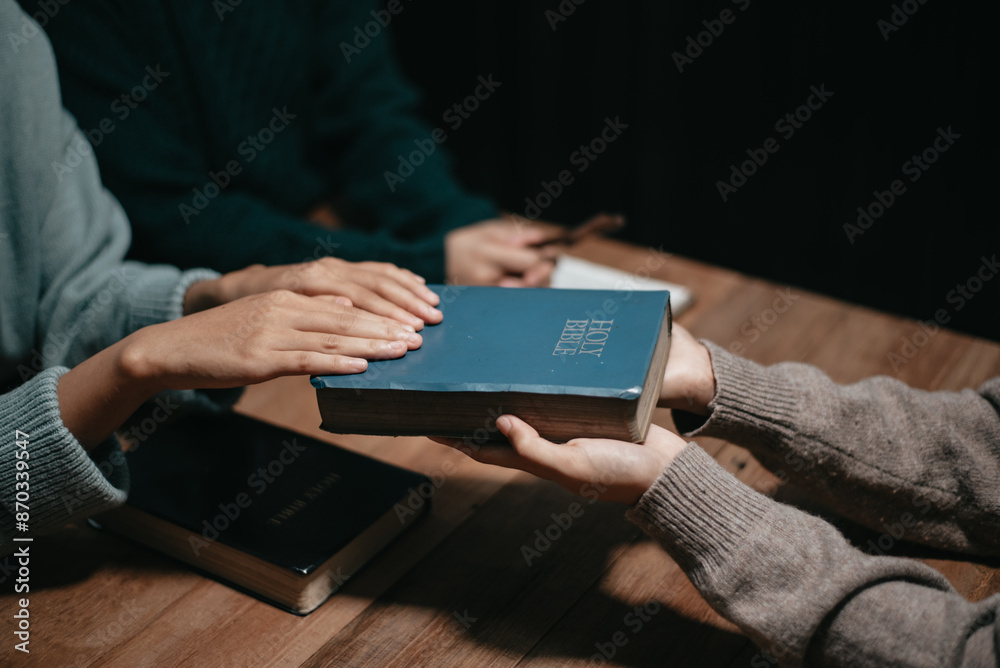 Group of Christians sit together and pray around a wooden table with blurred open Bible pages in their homeroom. Group of Christians sit together and pray around a wooden table with blurred open Bible pages in their homeroom.