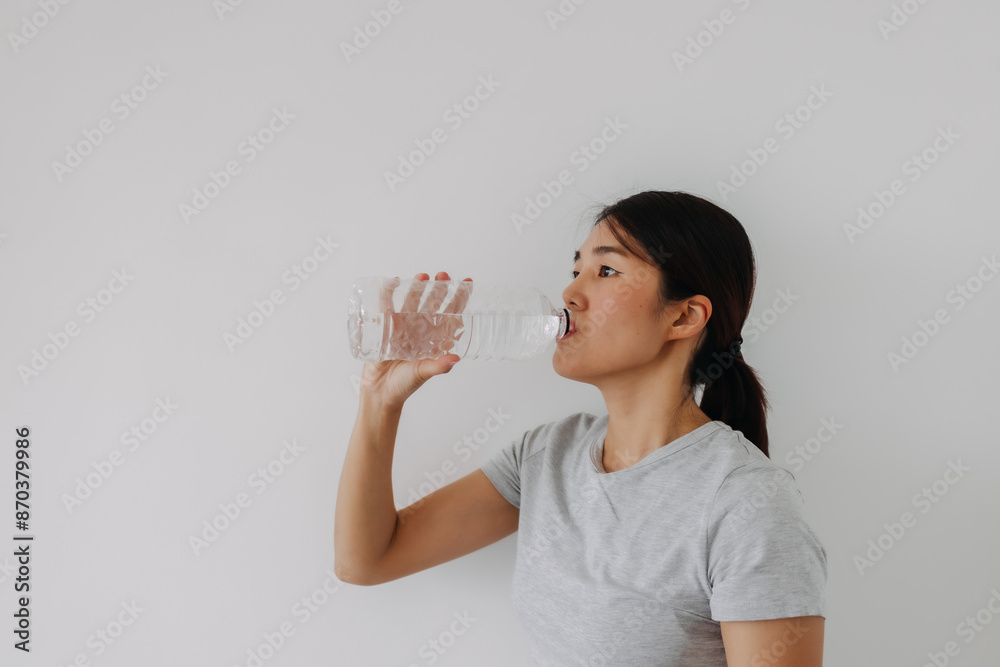 Side view of asian Thai woman drinking mineral water from the bottle, standing isolated over white background wall. Side view of asian Thai woman drinking mineral water from the bottle, standing isolated over white background wall.