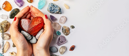 A female hand offers a heart-shaped crystal surrounded by various healing stones on a white background, creating a serene copy space image. photo