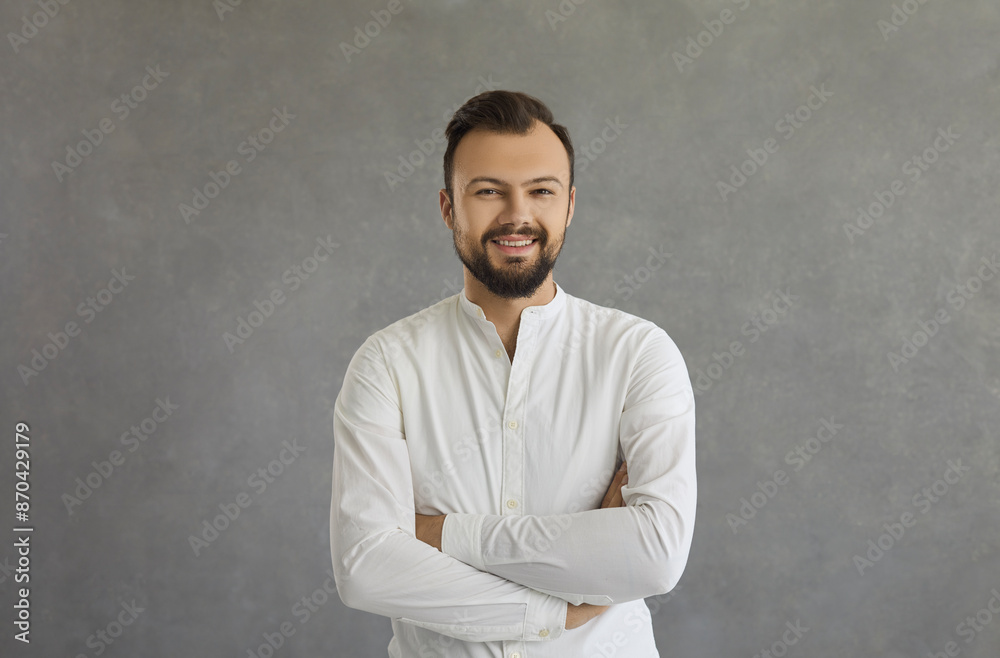 Portrait of a handsome happy caucasian man standing with arms crossed on gray background. Bearded millennial man in white shirt smiling looking at camera. Concept of confident and successful men. Portrait of a handsome happy caucasian man standing with arms crossed on gray background. Bearded millennial man in white shirt smiling looking at camera. Concept of confident and successful men.