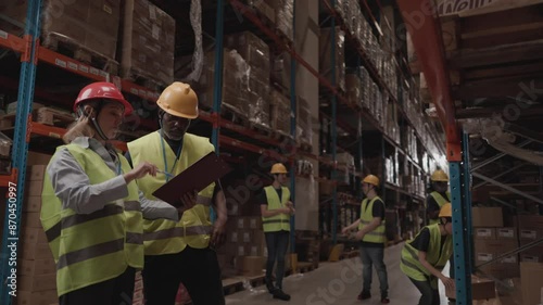 Team of multiracial warehouse workers, wearing security gear discussing inventory while checking clipboard. Busy warehouse environment. Warehouse workers discussing inventory