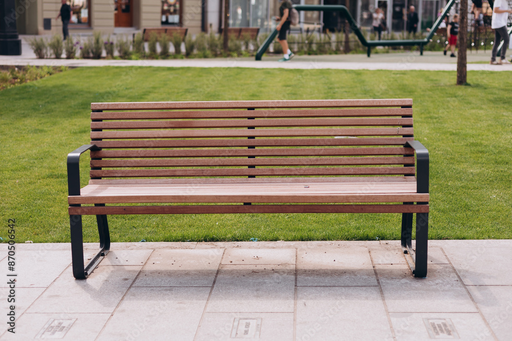 A modern wooden bench standing in the park by the sidewalk. Comfortable bench in recreation area. City improvement, urban planning, public spaces. A modern wooden bench standing in the park by the sidewalk. Comfortable bench in recreation area. City improvement, urban planning, public spaces.