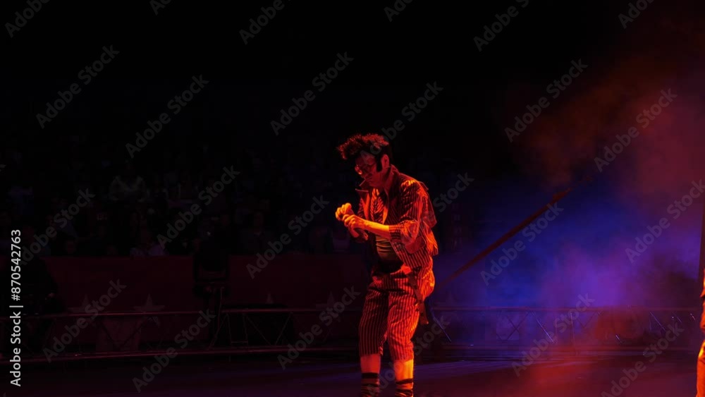 A clown with a funny hairstyle entertains guests in the circus arena in the light of colorful spotlights and smoke. He's tap dancing and singing into a microphone. Circus performance. A clown with a funny hairstyle entertains guests in the circus arena in the light of colorful spotlights and smoke. He's tap dancing and singing into a microphone. Circus performance.