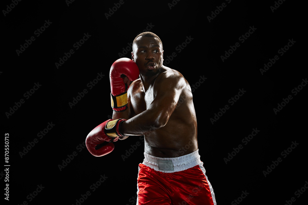 Fototapeta premium Dynamic image of serious African-American man, boxer with muscular boy, in red gloves and short punching, training isolated on black background. Concept of sport, active lifestyle, body, strength