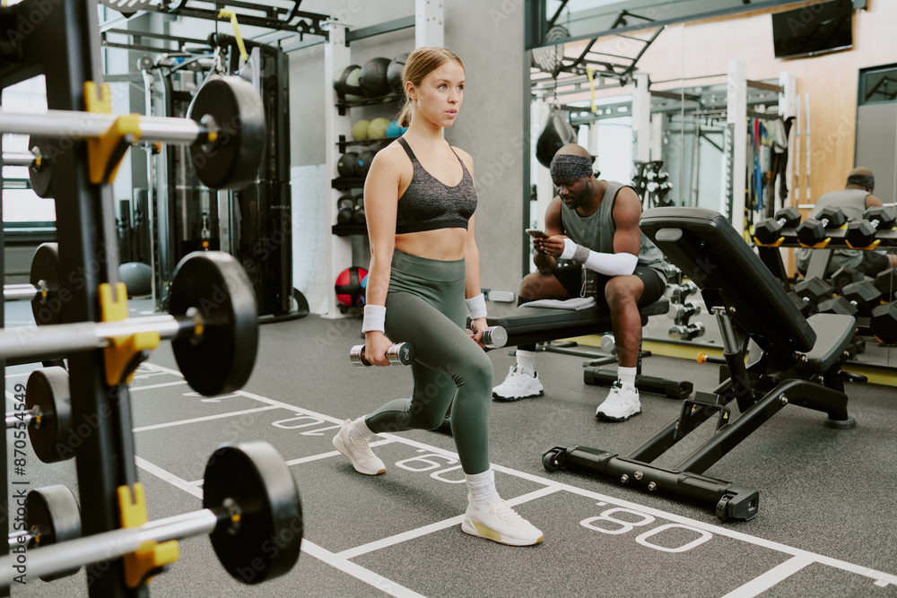 Newbie doing barbell lunge while her African American male coach sitting on bench and writing her training routine in phone Newbie doing barbell lunge while her African American male coach sitting on bench and writing her training routine in phone