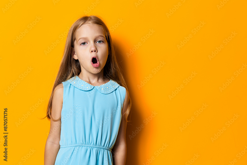 Joyful little girl in blue dress expressing excitement with wide open mouth against vibrant orange background Joyful little girl in blue dress expressing excitement with wide open mouth against vibrant orange background